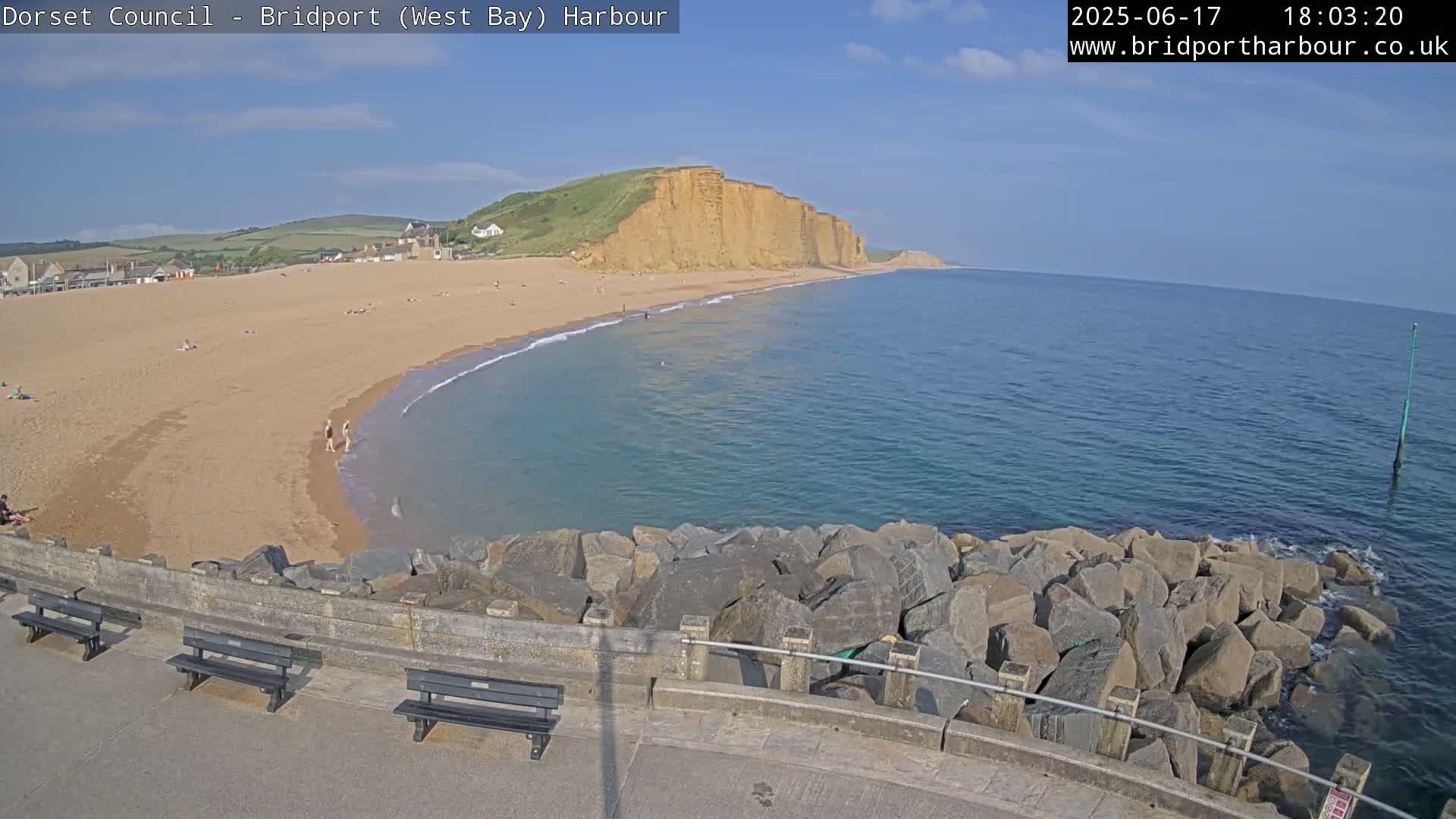 A sandy beach with people on it curves around a calm, blue ocean bordered by a rocky breakwater and high, tan cliffs under a mostly clear sky.