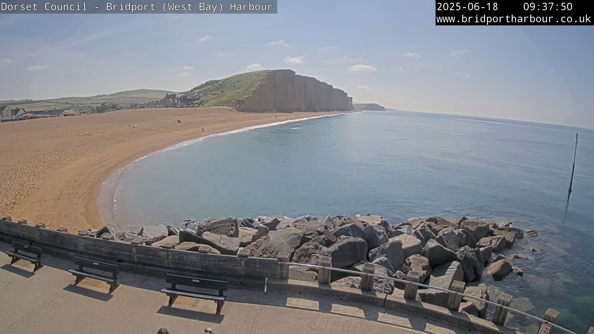 A sandy beach curves into a calm sea under a sunny sky, with a rocky breakwater and benches in the foreground.