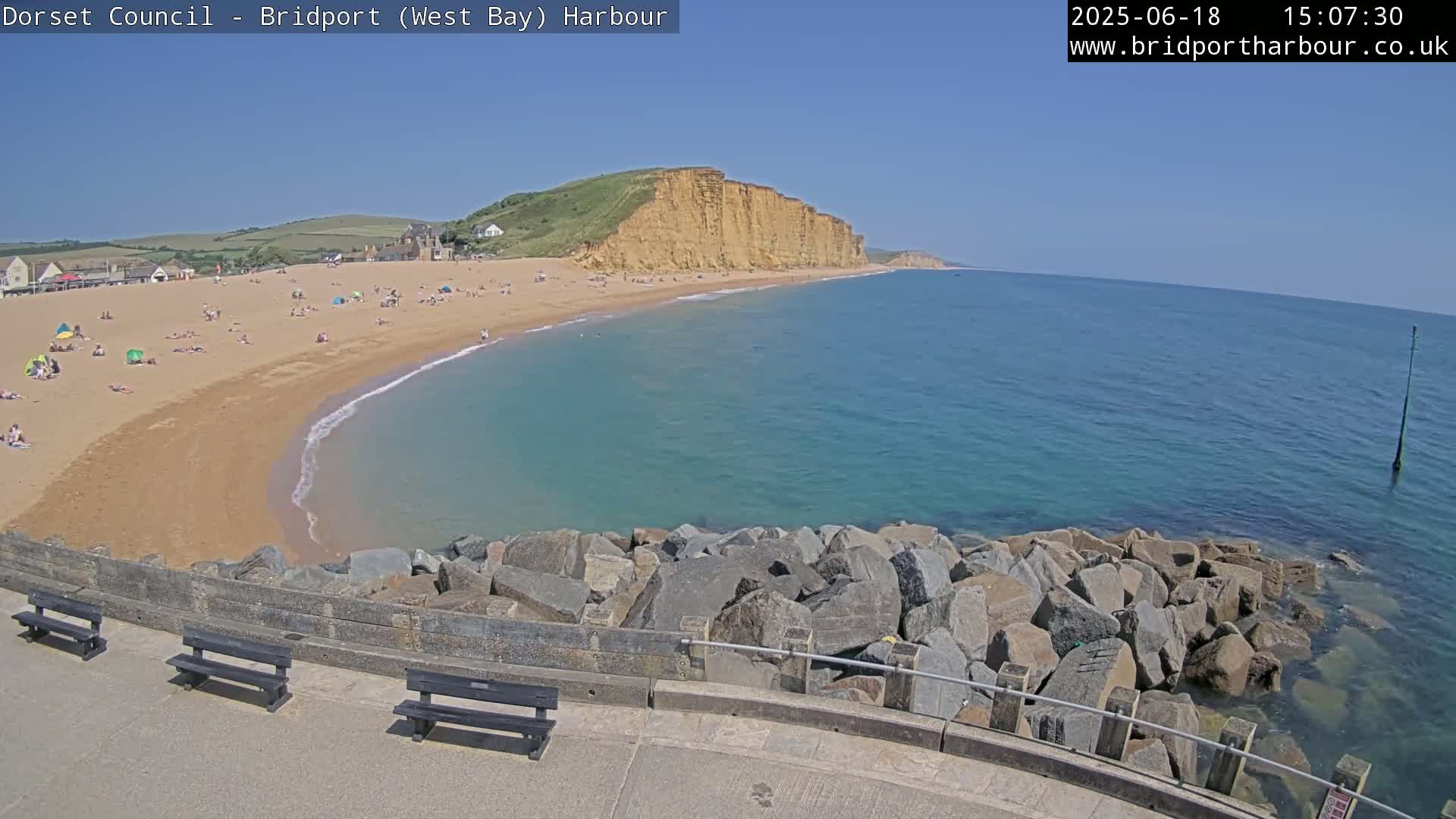 A sunny day at a sandy beach with calm, clear turquoise water, backed by high cliffs and a rock breakwater in the foreground.