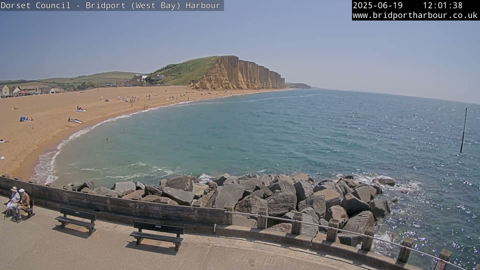 A sandy beach with people sunbathing and swimming is situated below a line of cliffs, all overlooking a calm, blue ocean under a clear sky.