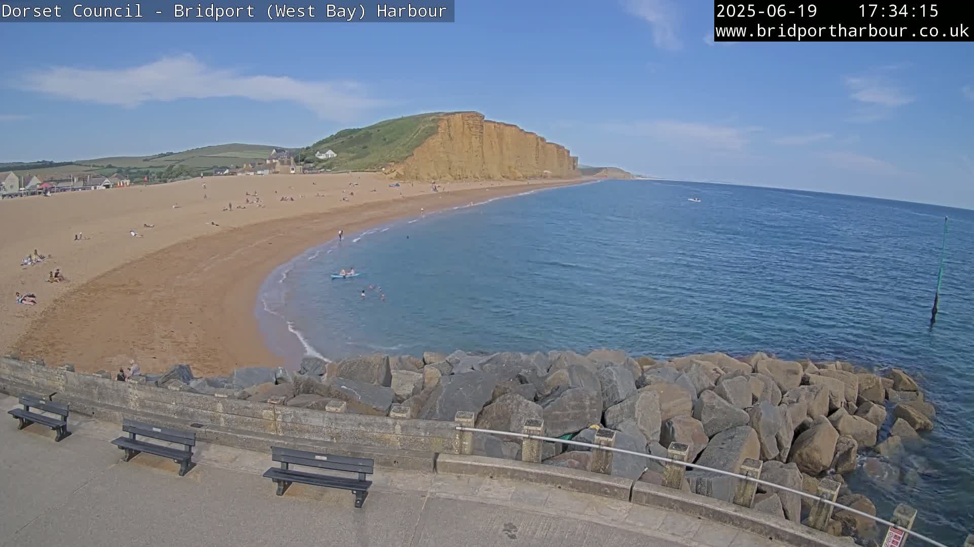 A sunny day at a sandy beach with calm, blue water,  tall cliffs in the background, and a rocky breakwater in the foreground.