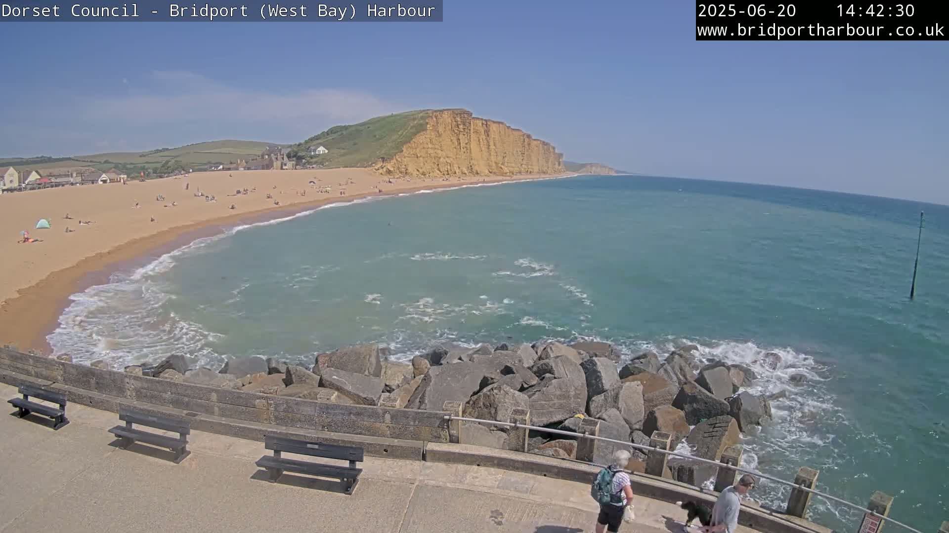 A sunny day at a beach with a sandy shoreline, a rocky breakwater, and a large cliff face in the background.