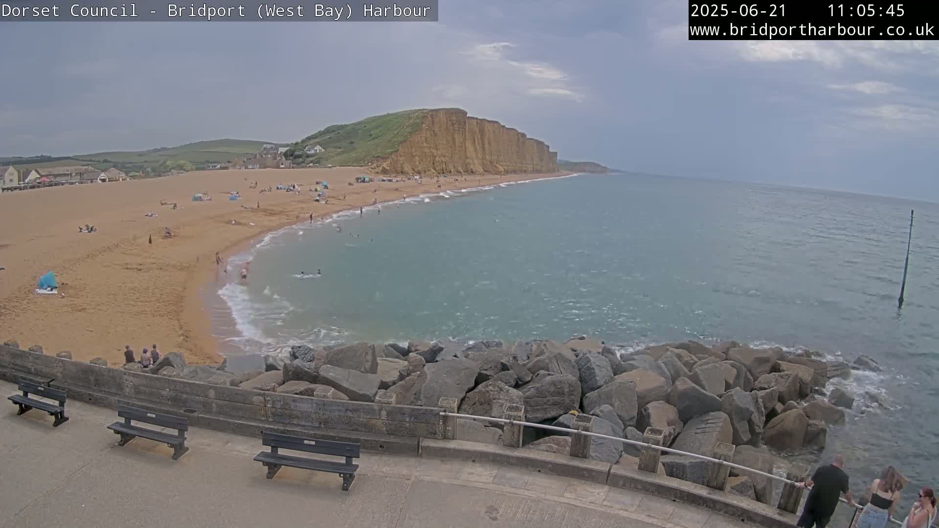 A sandy beach with people on it is next to a calm, blue ocean that meets a rocky breakwater under a partly cloudy sky.