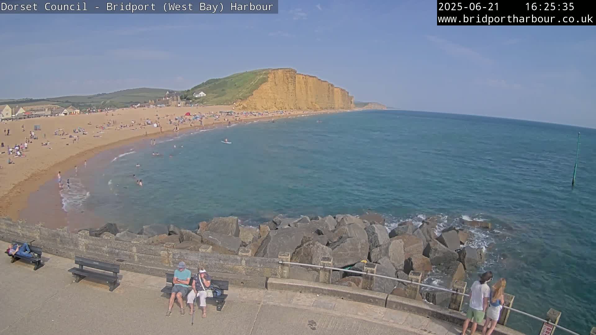 A sunny day at a crowded beach with calm, clear blue water, backed by a high, sandy cliff.
