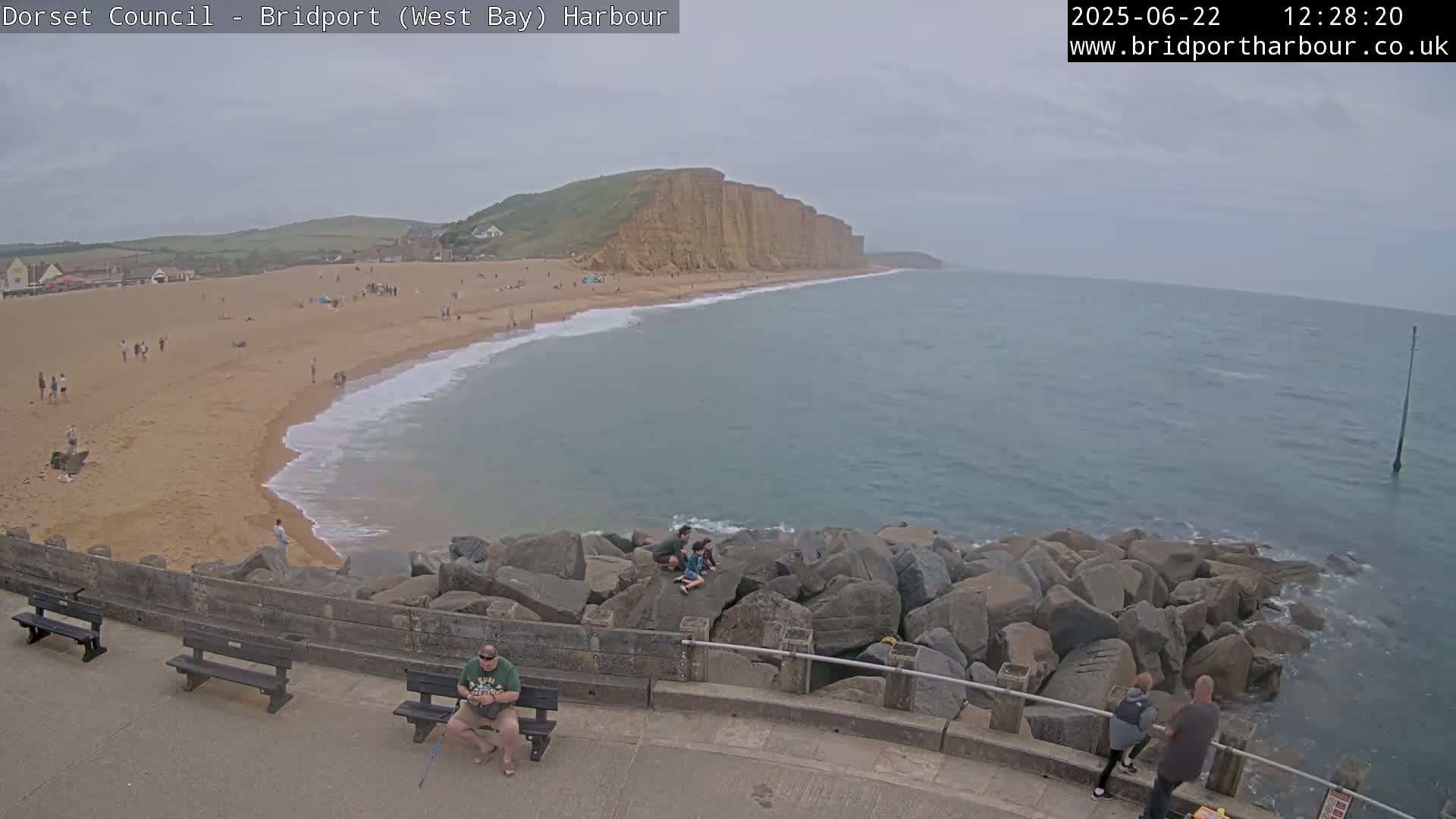 A sandy beach with many people on it curves around a rocky point into a calm, grey-blue sea under an overcast sky.