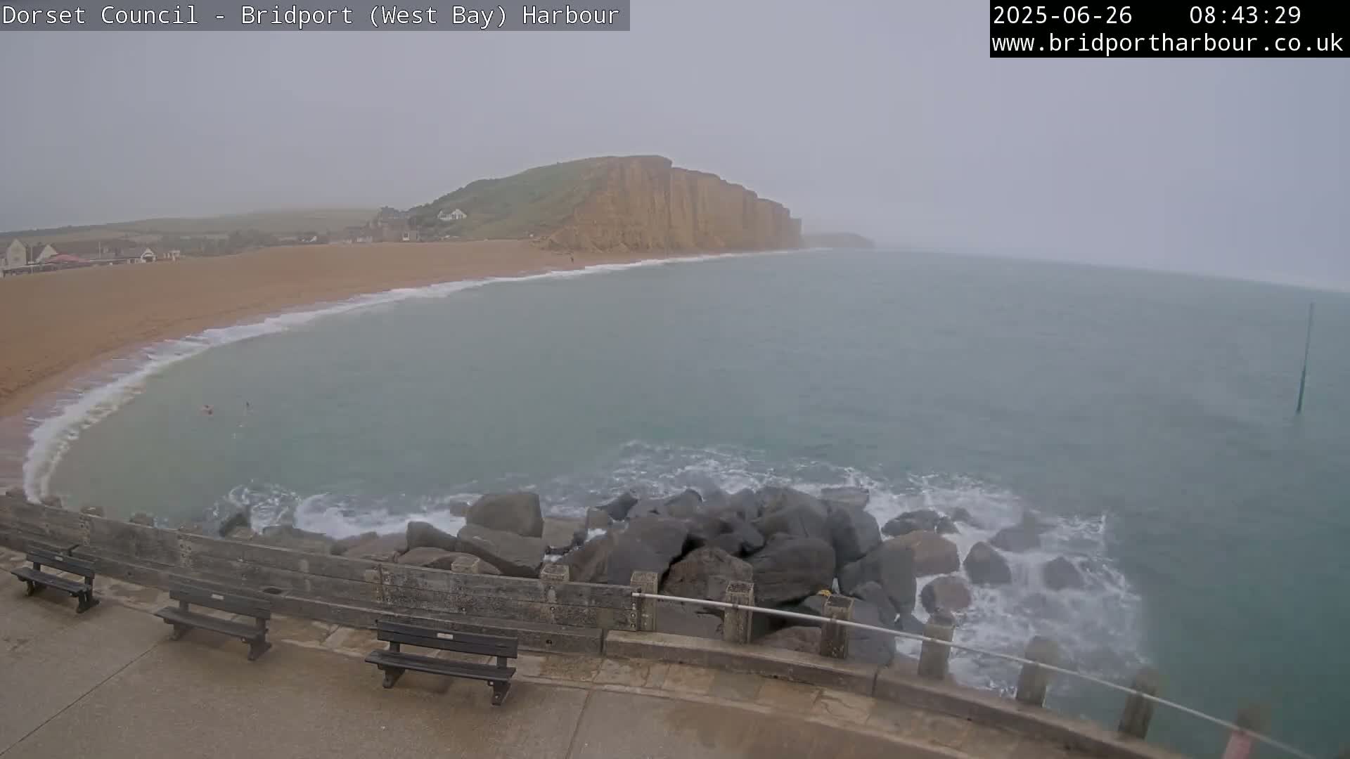A hazy, overcast day shows a sandy beach curving into a bay, with dark rocks and waves crashing against a sea wall in the foreground and cliffs visible in the distance.