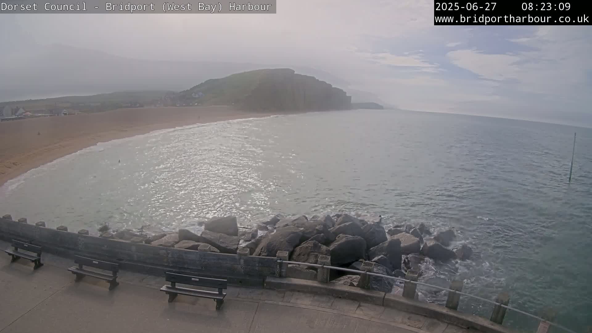 A hazy view shows a sandy beach curving into a calm sea lapping against a rocky breakwater, with benches on a paved walkway.