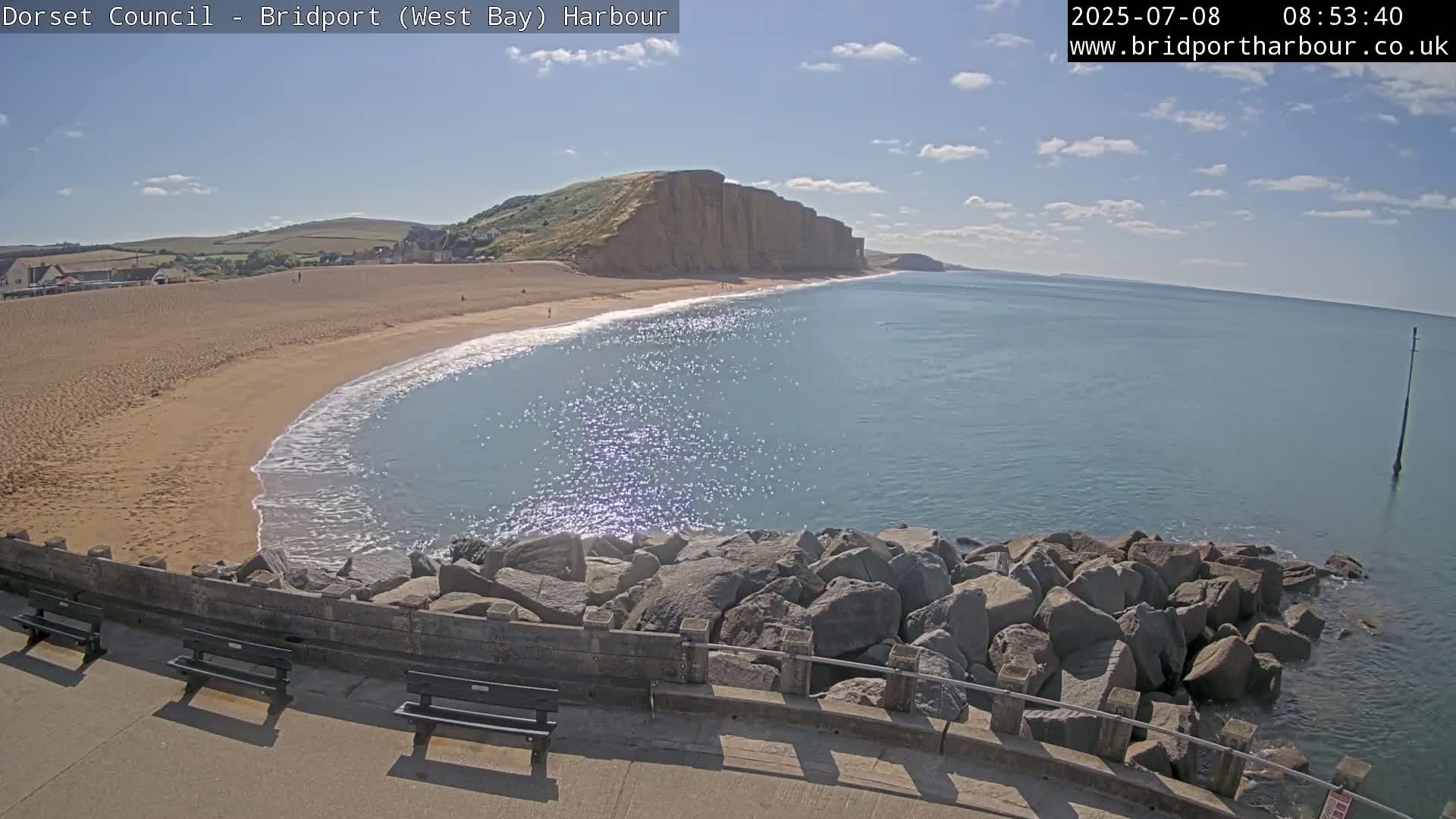 A sandy beach curves around a calm, sunlit ocean, backed by a high cliff face under a mostly clear sky.