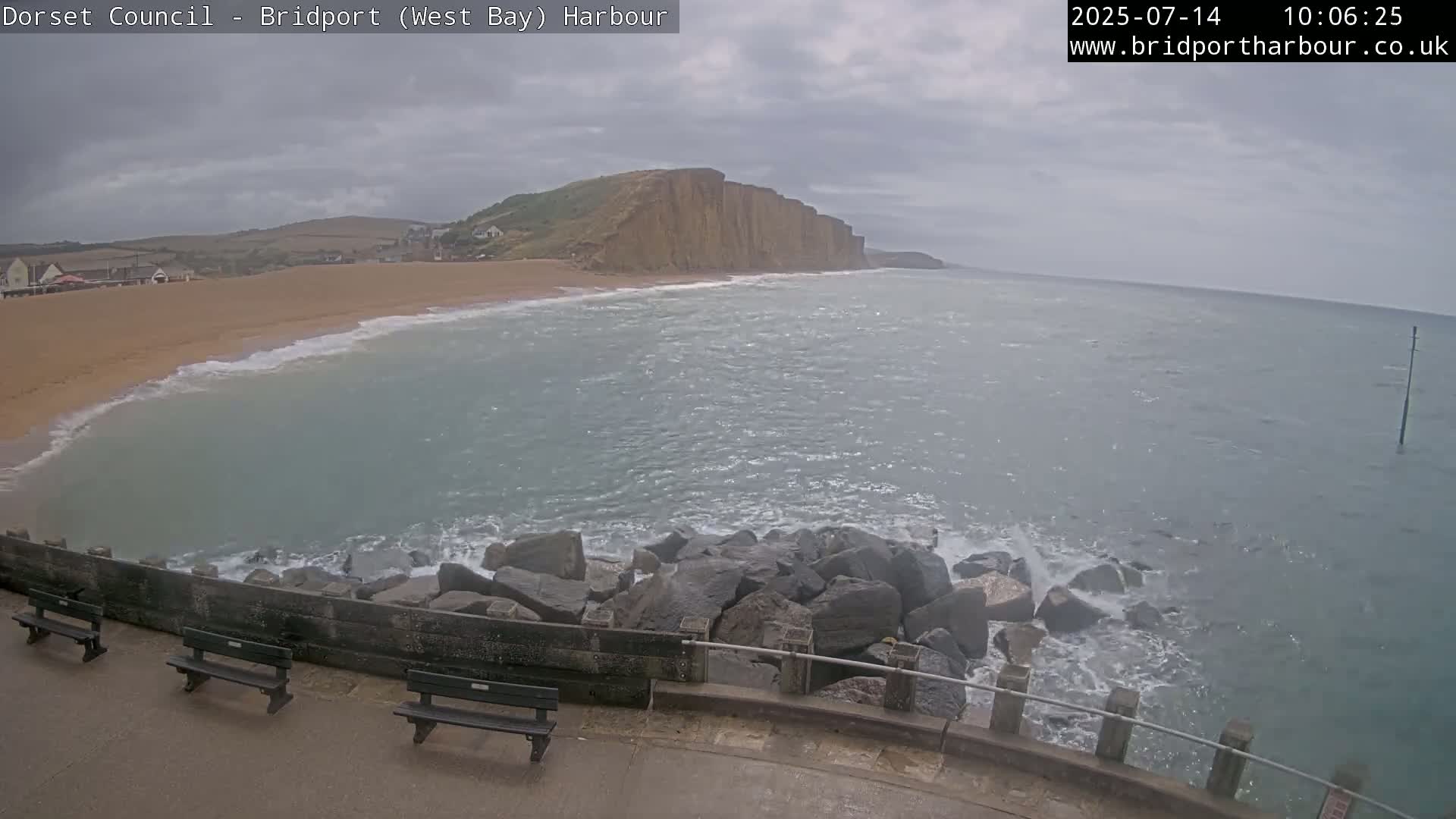 A view from a harbor shows a sandy beach, a large cliff face, and a choppy sea under an overcast sky.