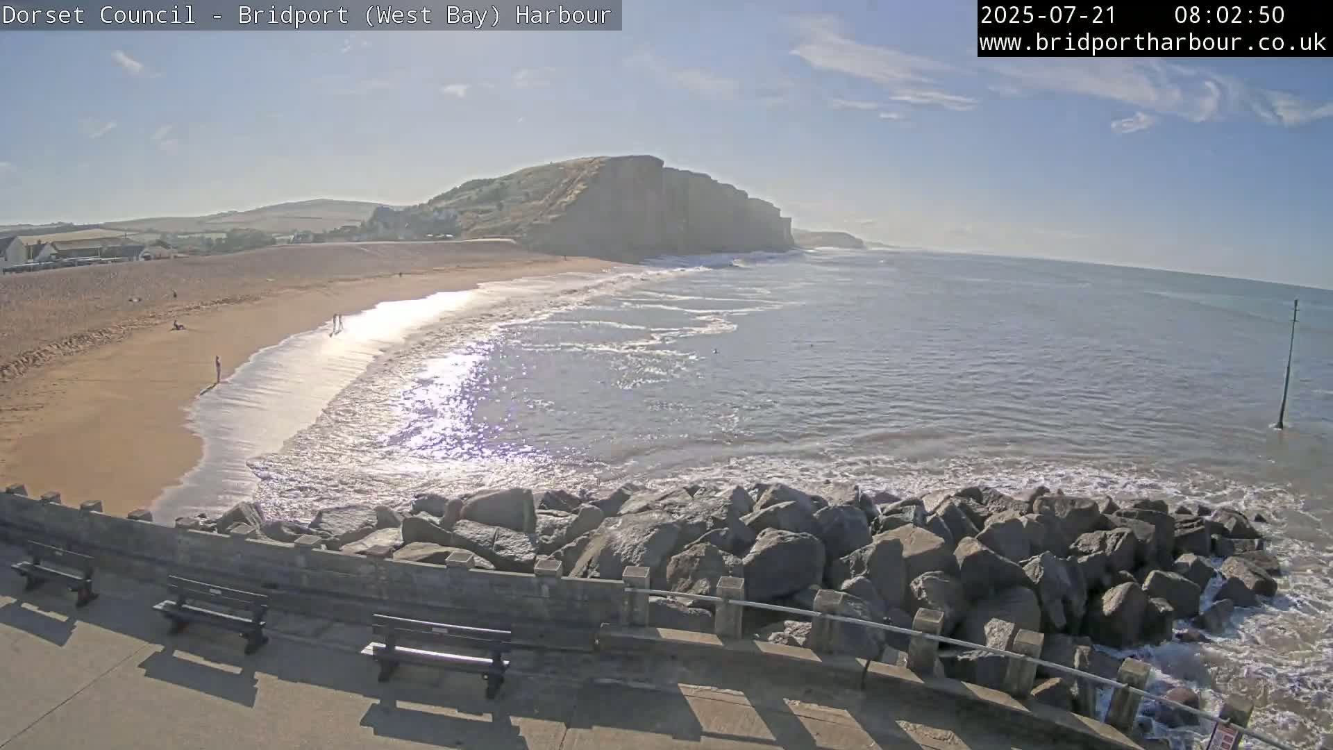A sandy beach with a few people on it is next to a calm sea with small waves lapping against a rocky breakwater under a mostly sunny sky.