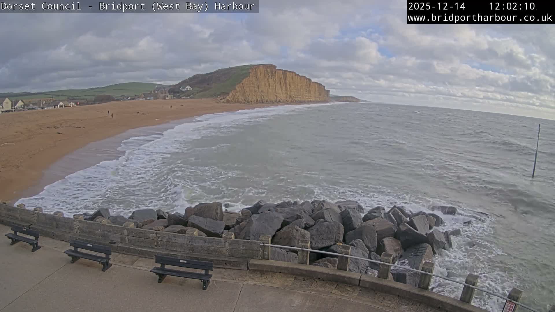 A wide coastal view shows a sandy beach with breaking waves, backed by green hills, a town, and towering cliffs, all under an overcast sky with a faint glow on the horizon, observed from a promenade with benches and a rocky sea defense.