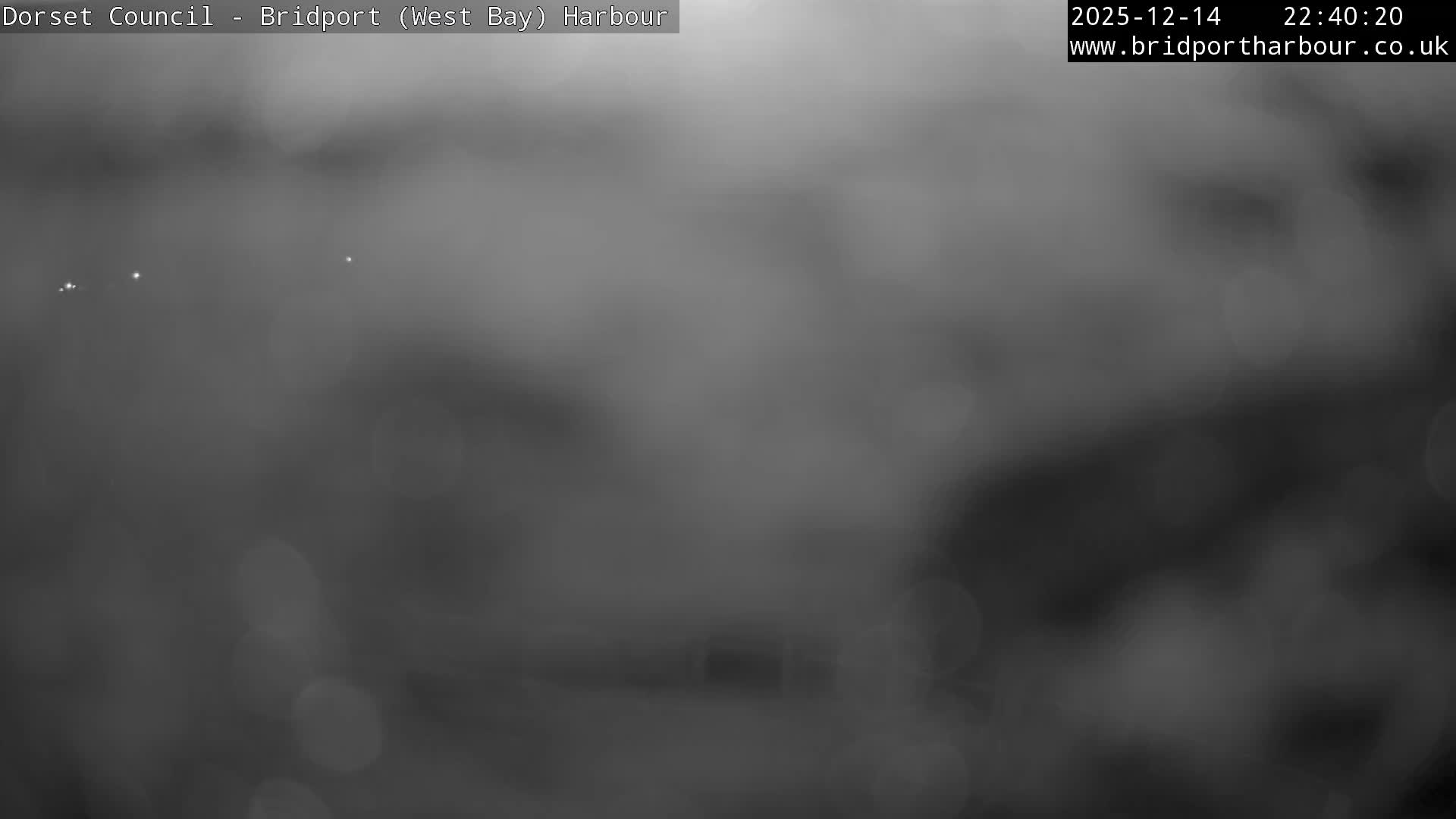 A wide coastal view shows a sandy beach with breaking waves, backed by green hills, a town, and towering cliffs, all under an overcast sky with a faint glow on the horizon, observed from a promenade with benches and a rocky sea defense.