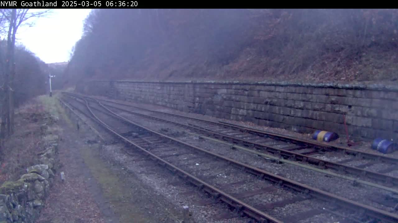 A misty view of a railway track curving through a stone-walled cutting, with two barrels lying beside the rails.