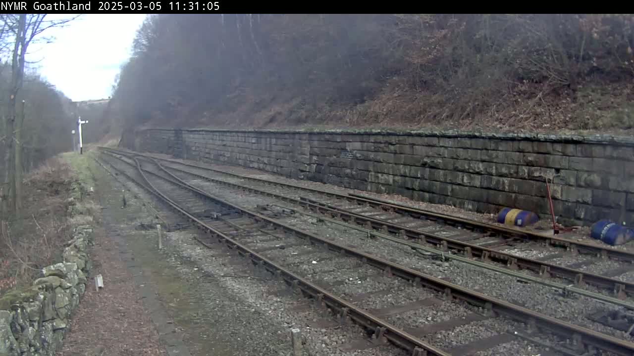 Two sets of train tracks curve gently to the right, alongside a stone retaining wall, in an overcast outdoor setting.