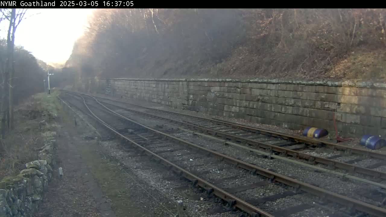 A misty view of railway tracks curving alongside a stone retaining wall, bordered by trees and vegetation.