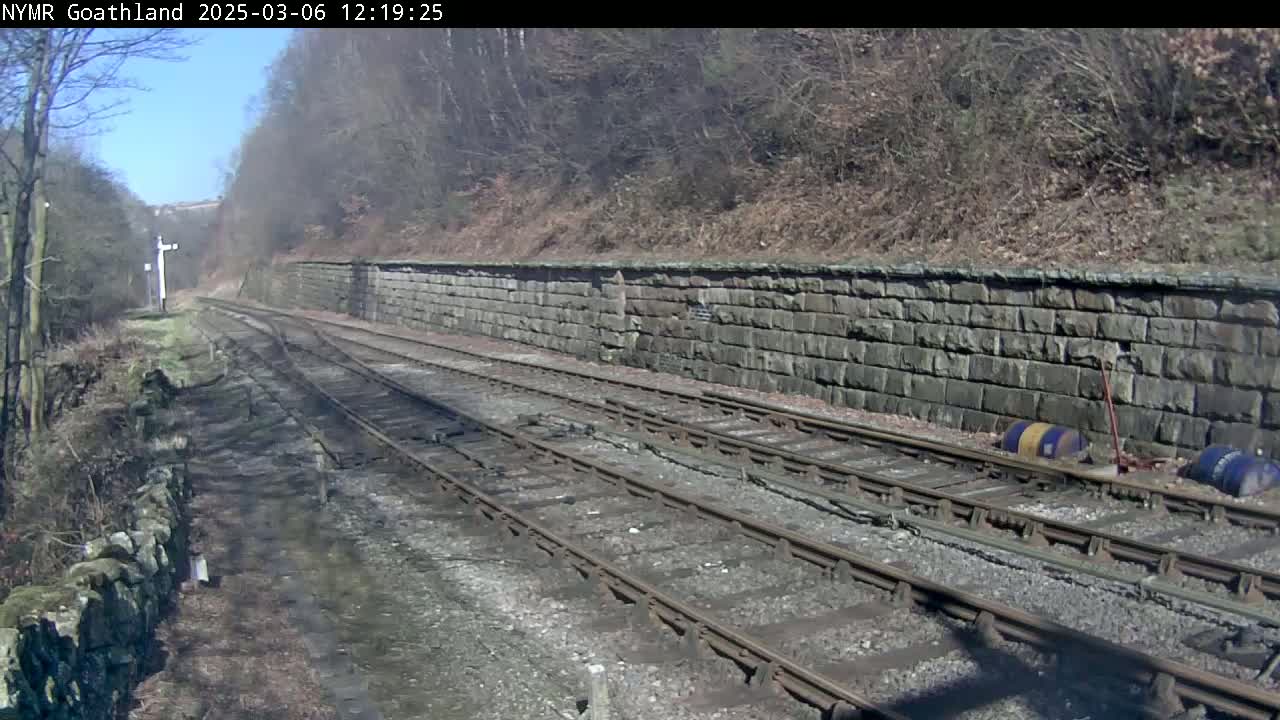 A sunny day reveals a view of railway tracks bordered by a stone wall and a wooded hillside.