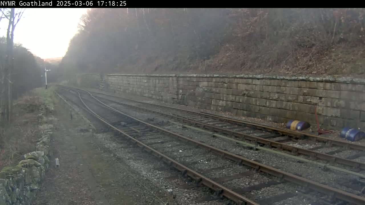 A foggy view of a railway track and stone retaining wall alongside a wooded hillside.