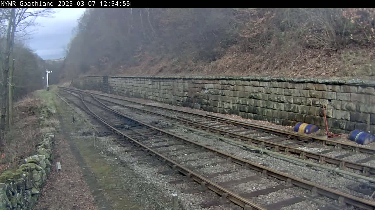 A view of train tracks curving alongside a stone wall, under an overcast sky, with bare winter trees and shrubs in the background.