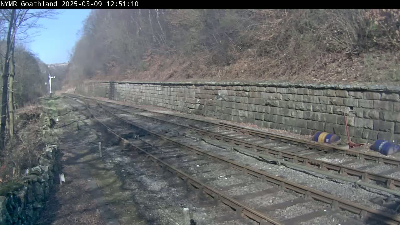 A sunny day reveals a railway track bordered by a stone wall and wooded hillside.