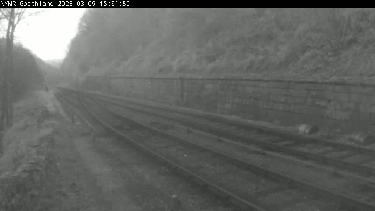 A misty overcast day reveals a view of dual railway tracks running alongside a low stone wall, with sparse vegetation visible on either side.