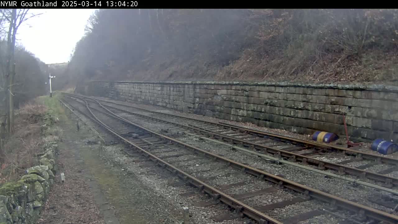 A misty view of train tracks curving alongside a stone wall, bordered by bare winter vegetation.