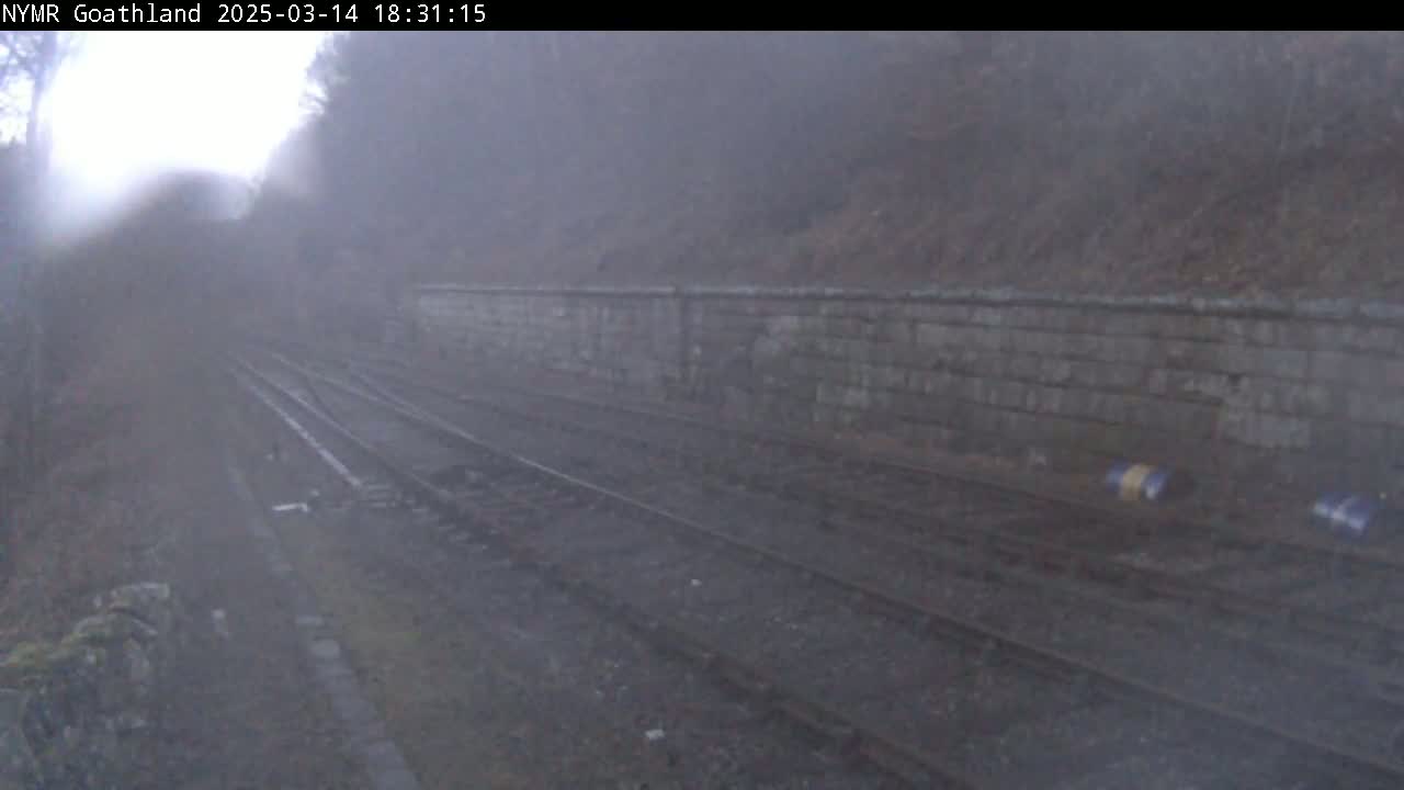 A foggy view of train tracks running alongside a stone wall in a wooded area.