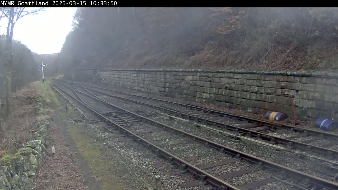 Several parallel train tracks extend into a foggy landscape alongside a low stone wall and sparse vegetation.