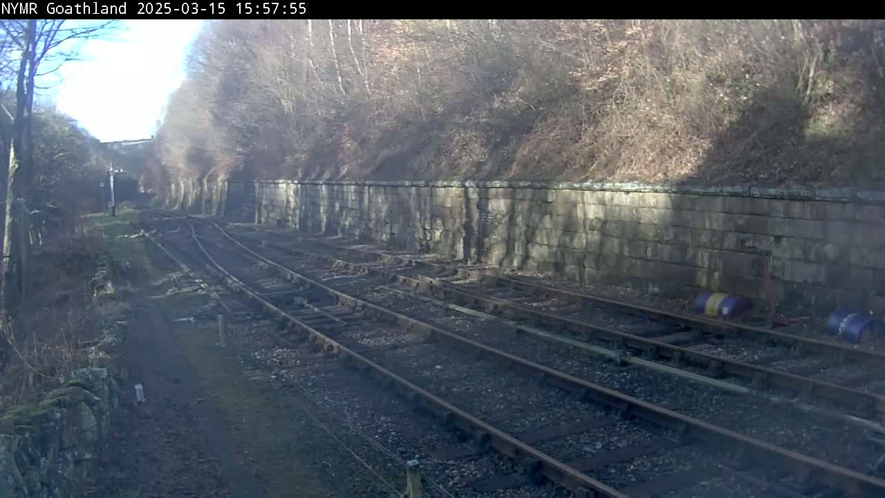 Multiple railway tracks run alongside a stone wall, bordered by leafless trees under a bright, sunny sky.