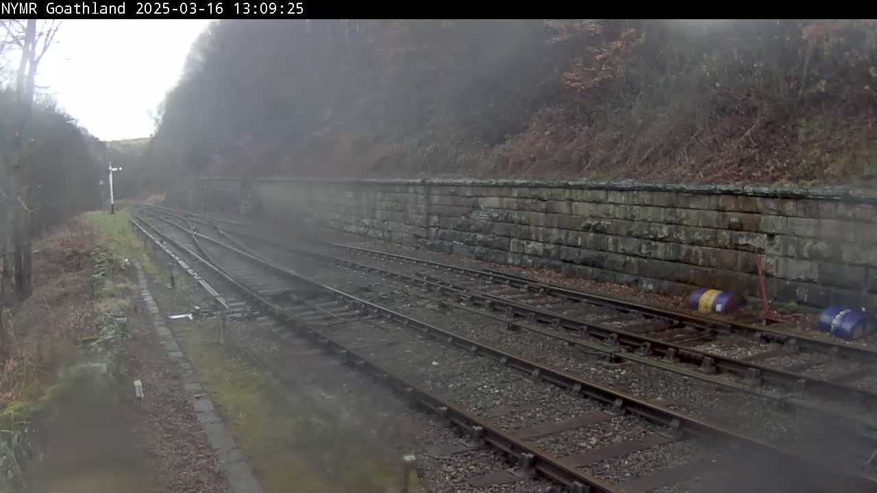 Train tracks curve through a foggy landscape alongside a low stone wall, with sparse vegetation and a few barrels visible near the tracks.