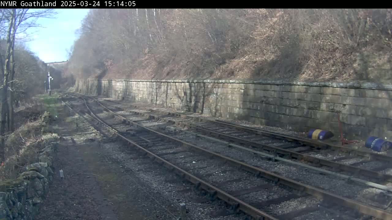 A sunny view of railway tracks alongside a stone retaining wall, bordered by leafless trees and shrubs.