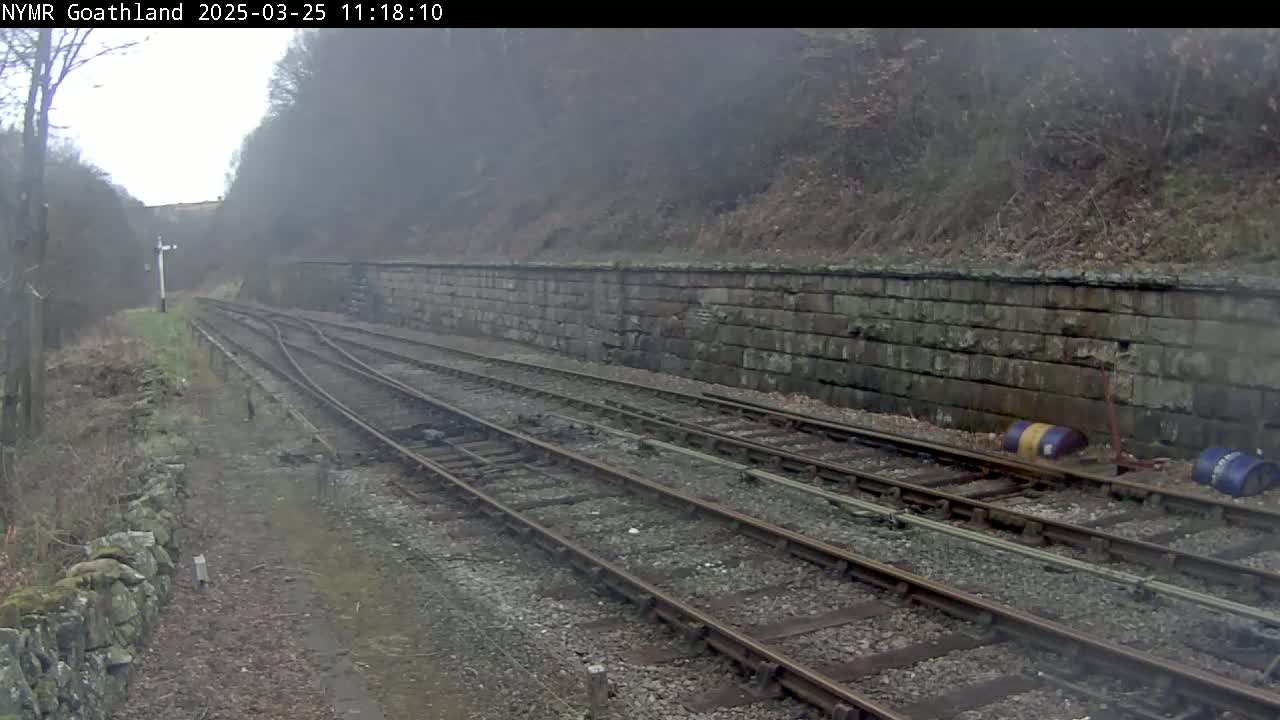A misty view of a railway track, curving slightly to the right, bordered by a low stone wall and woodland.