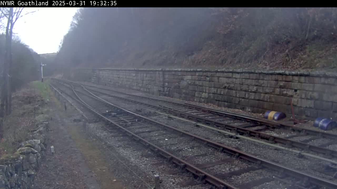 A misty view of railway tracks curving gently to the left, alongside a stone retaining wall and sparsely vegetated embankment.