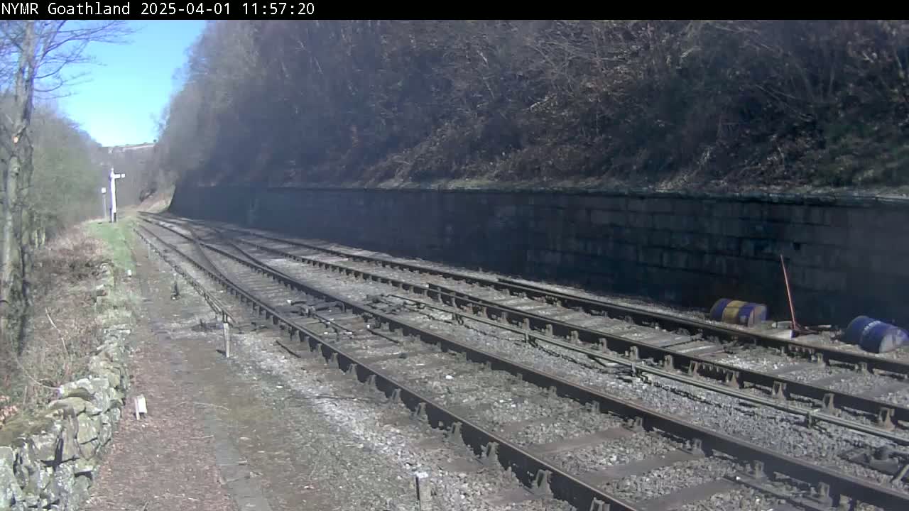 A sunny day shows a view of multiple train tracks alongside a stone wall and wooded hillside.