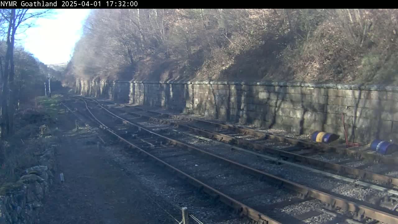 A sunny day reveals a railway track winding through a wooded area, alongside a stone retaining wall.