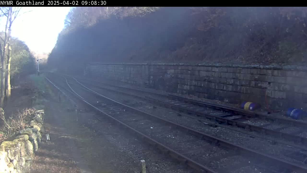 Three sets of train tracks curve gently to the right, alongside a stone wall, under a hazy, sunny sky.