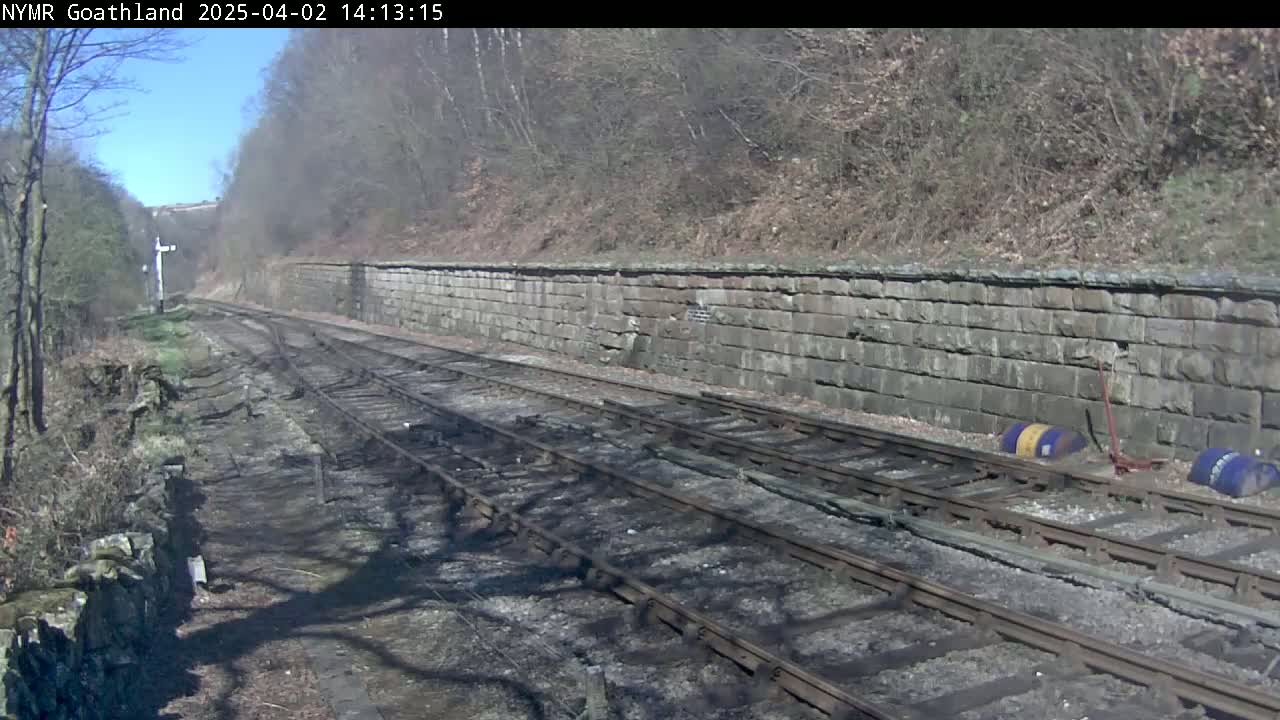 Under a sunny sky, train tracks curve gently to the right, flanked by a low stone wall and sparse vegetation.