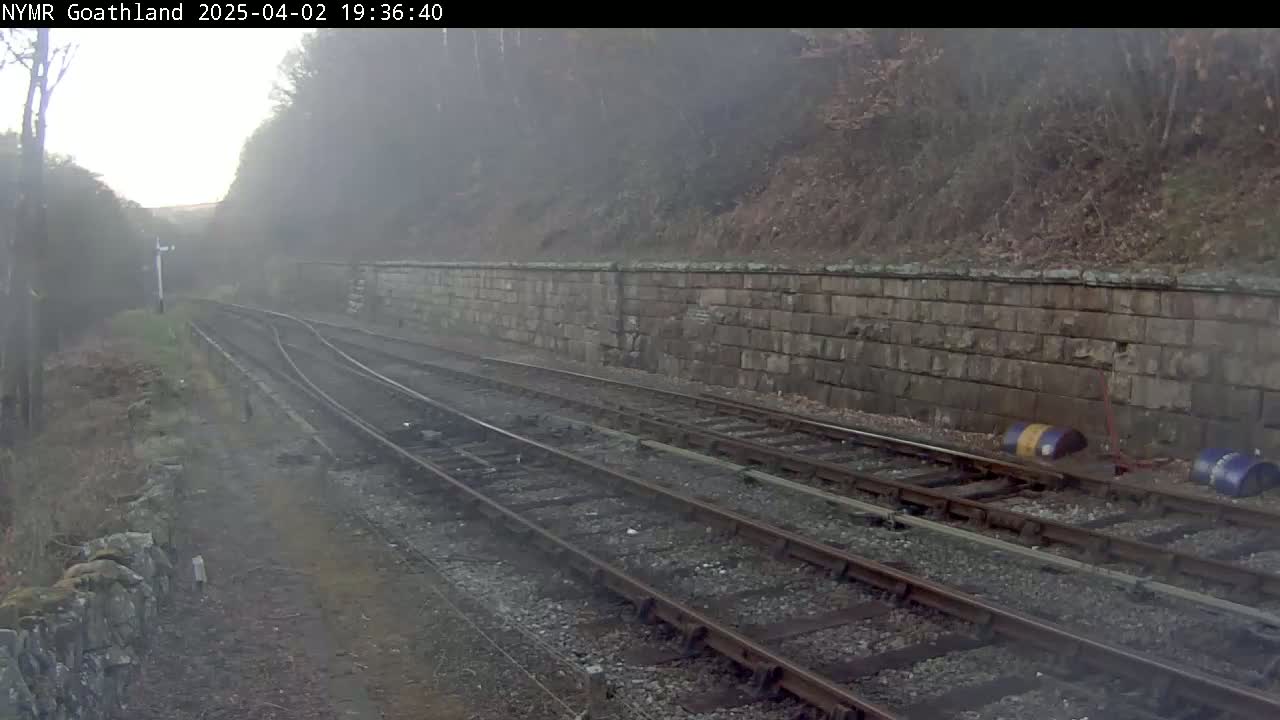 Two sets of train tracks curve through a foggy landscape alongside a low stone wall, with sparse vegetation visible.