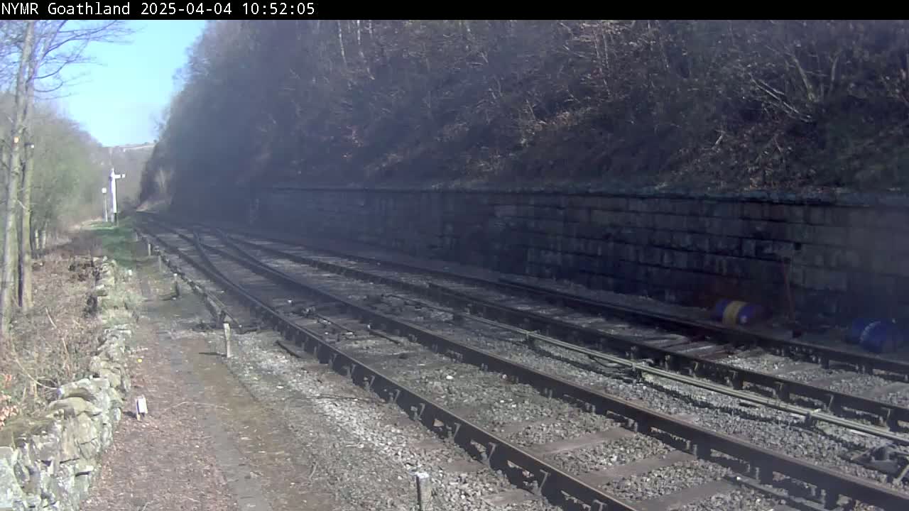 A sunny day reveals a railway track curving through a wooded area, alongside a stone wall.