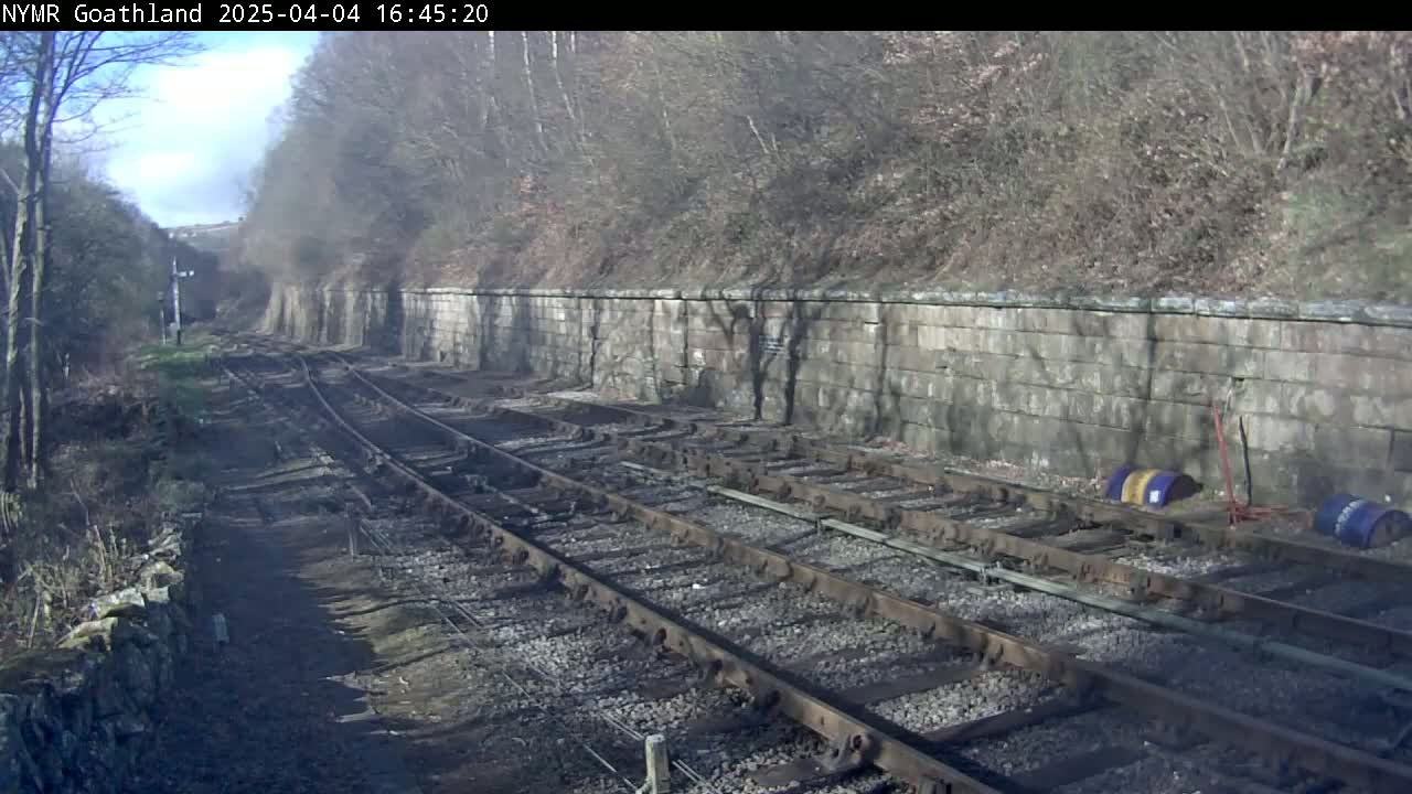 A sunny view of multiple railway tracks extending towards a stone wall and wooded hillside.