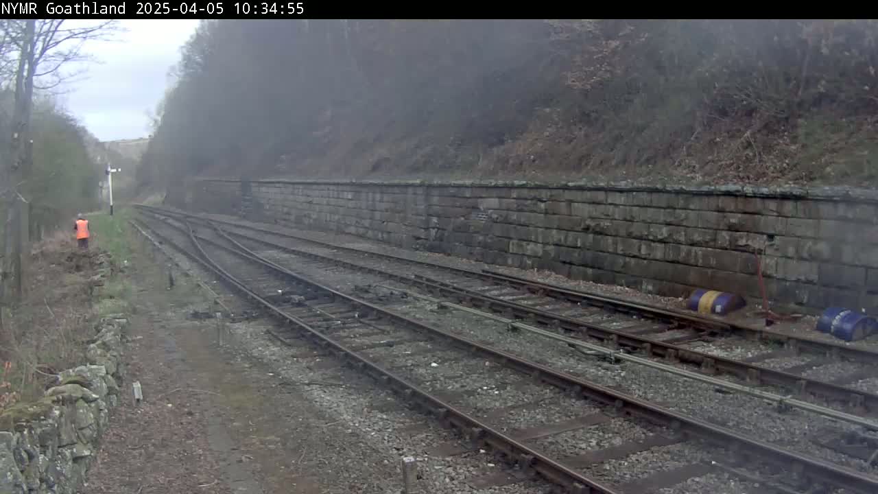 A foggy view of a railway track with a retaining wall alongside, a person in an orange vest visible in the distance.