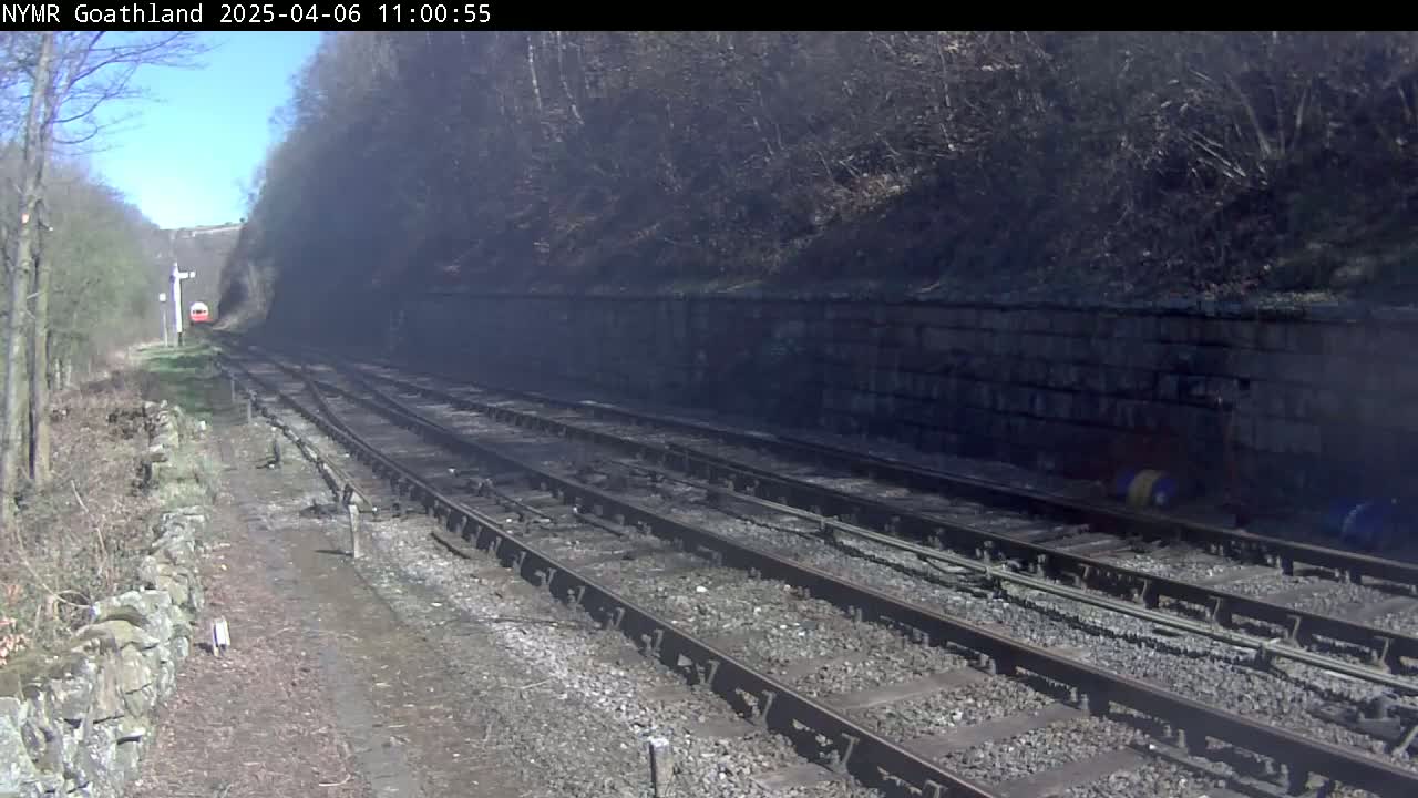 A red train is seen in the distance traveling along multiple train tracks that run beside a stone wall and into a wooded area on a sunny day.