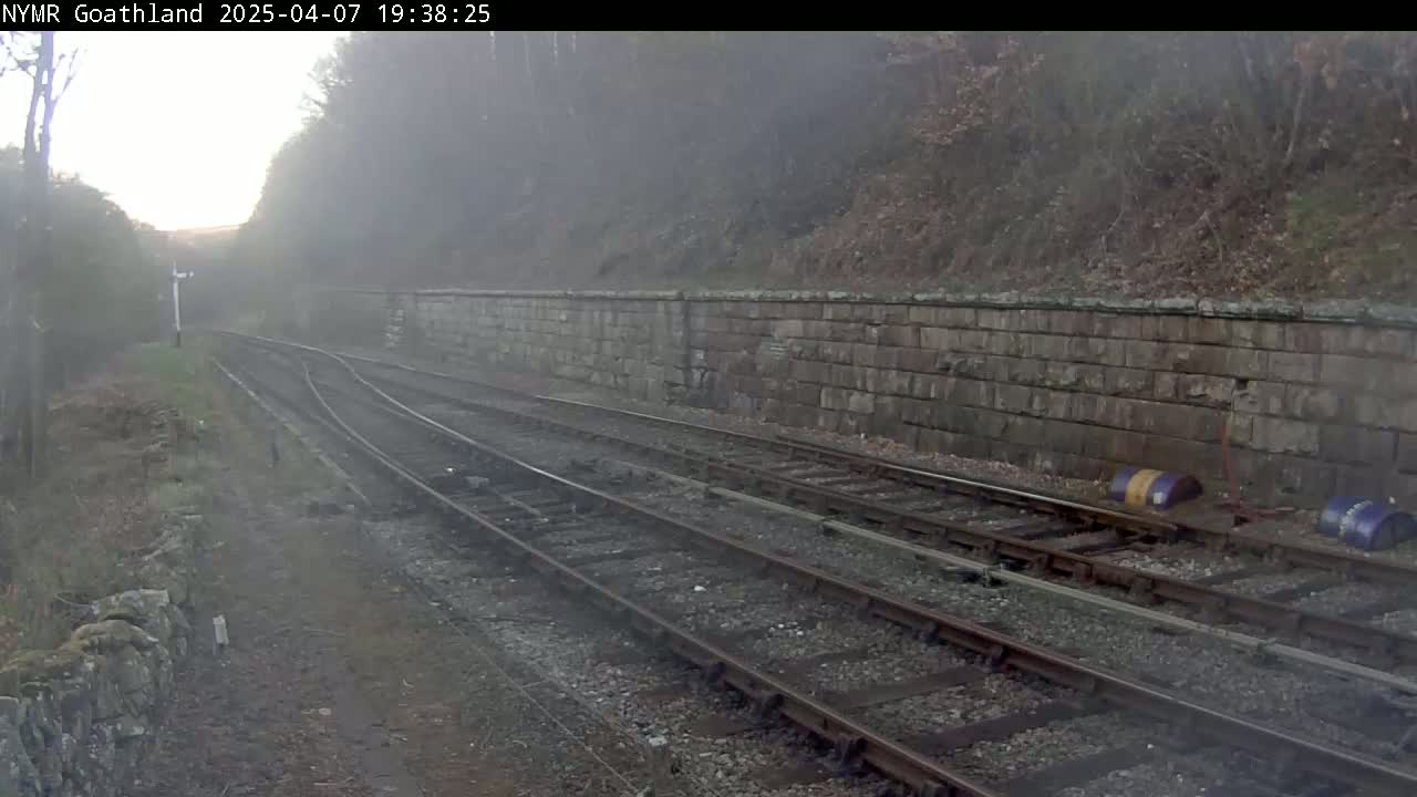 A foggy view of a railway line with a stone wall and some barrels next to the tracks.
