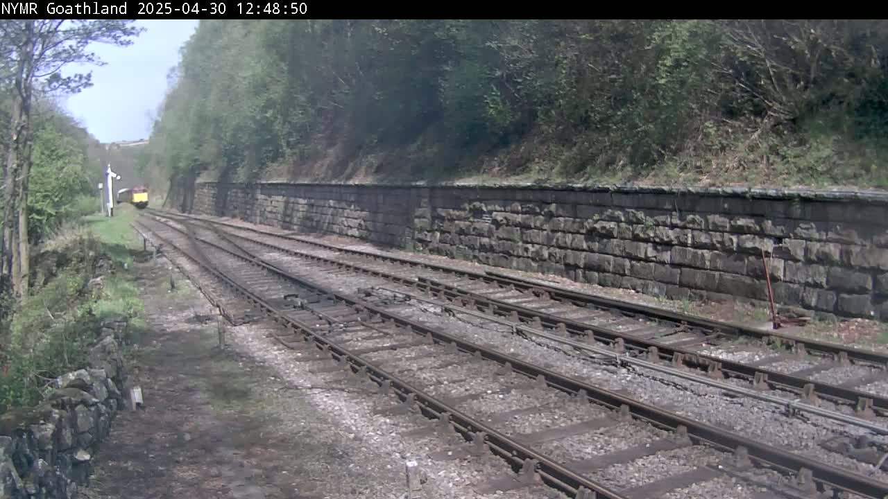 A yellow train is visible in the distance, traveling along a railway track that is flanked by a stone wall and lush green vegetation under a partly sunny sky.