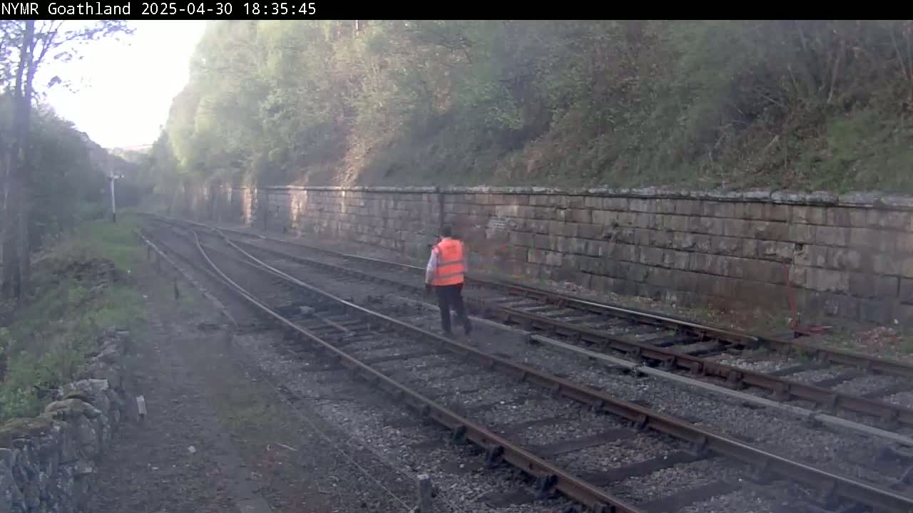 A person in a bright orange safety vest walks along railroad tracks on a hazy day, alongside a stone wall and lush greenery.