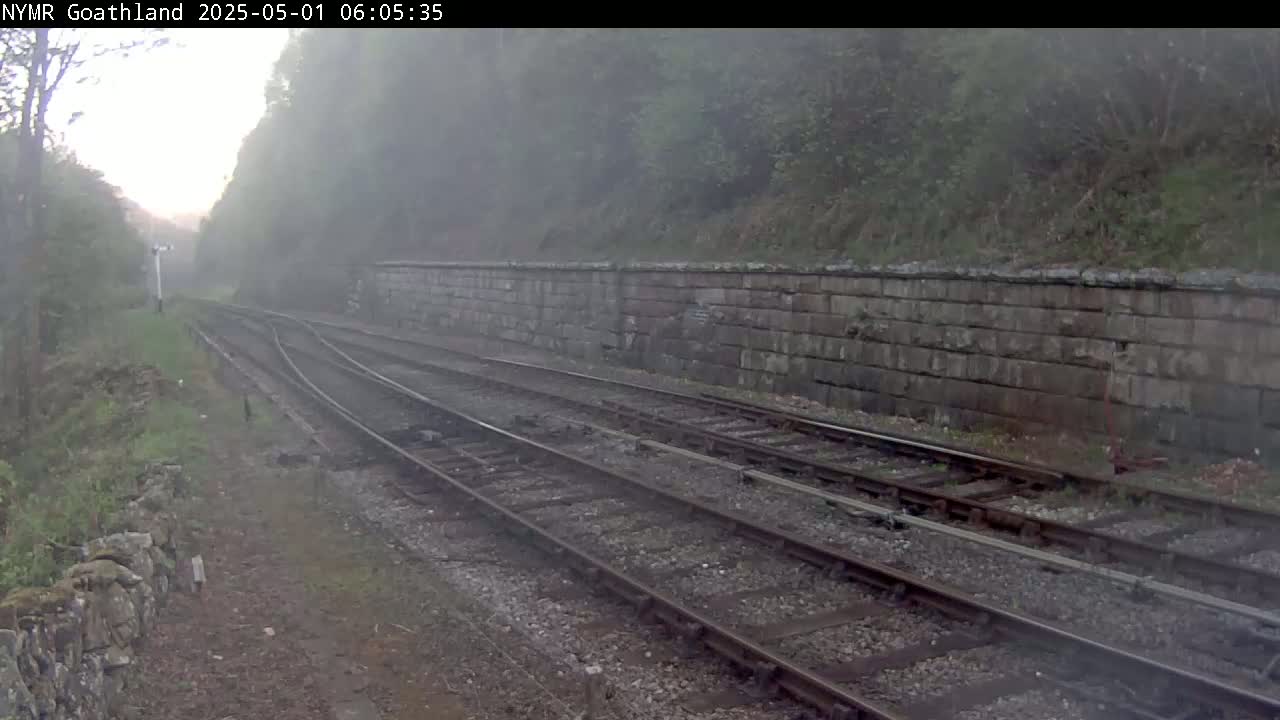 A foggy view of a railway track winding through a landscape with a stone retaining wall and lush green vegetation.