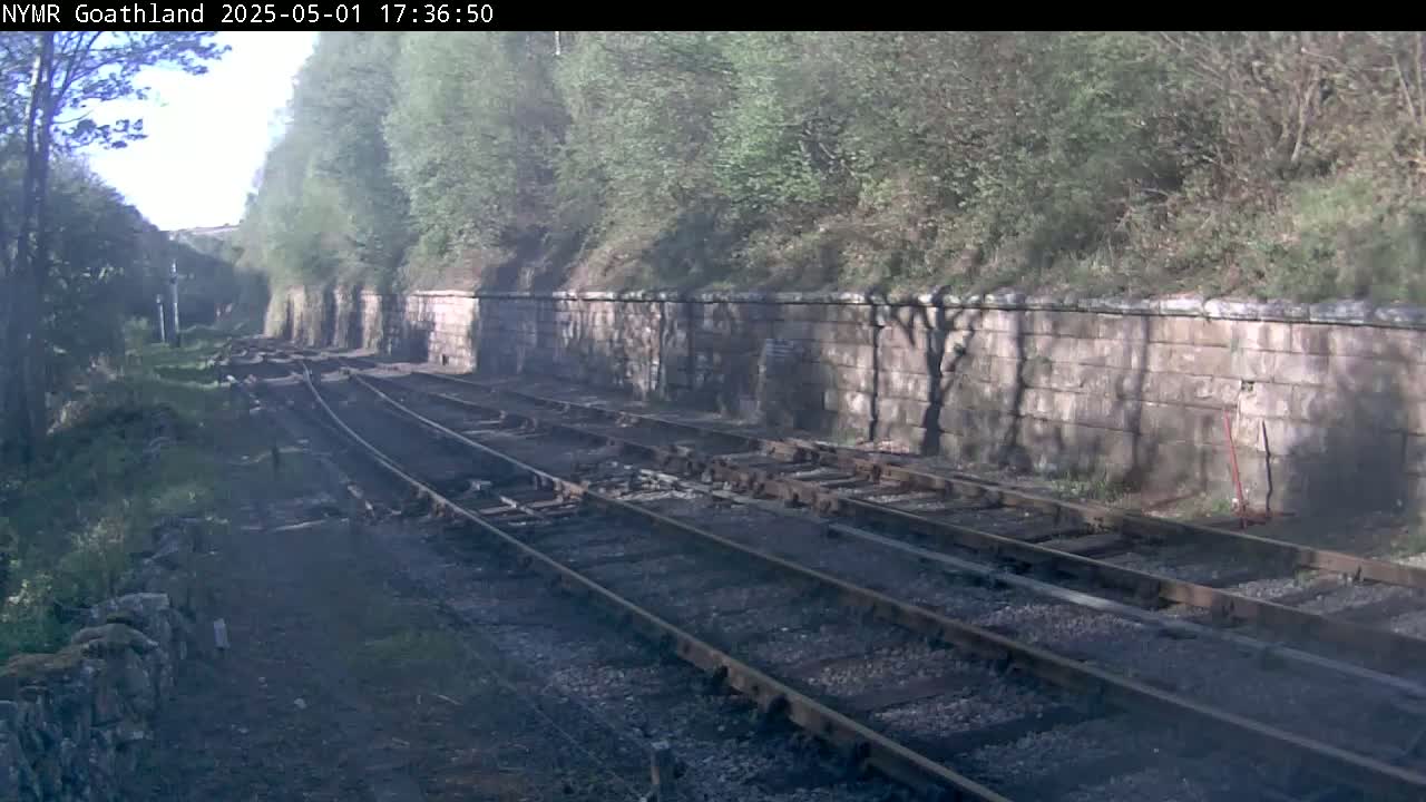 A view of railway tracks running alongside a stone wall, bordered by lush green trees under a partly sunny sky.