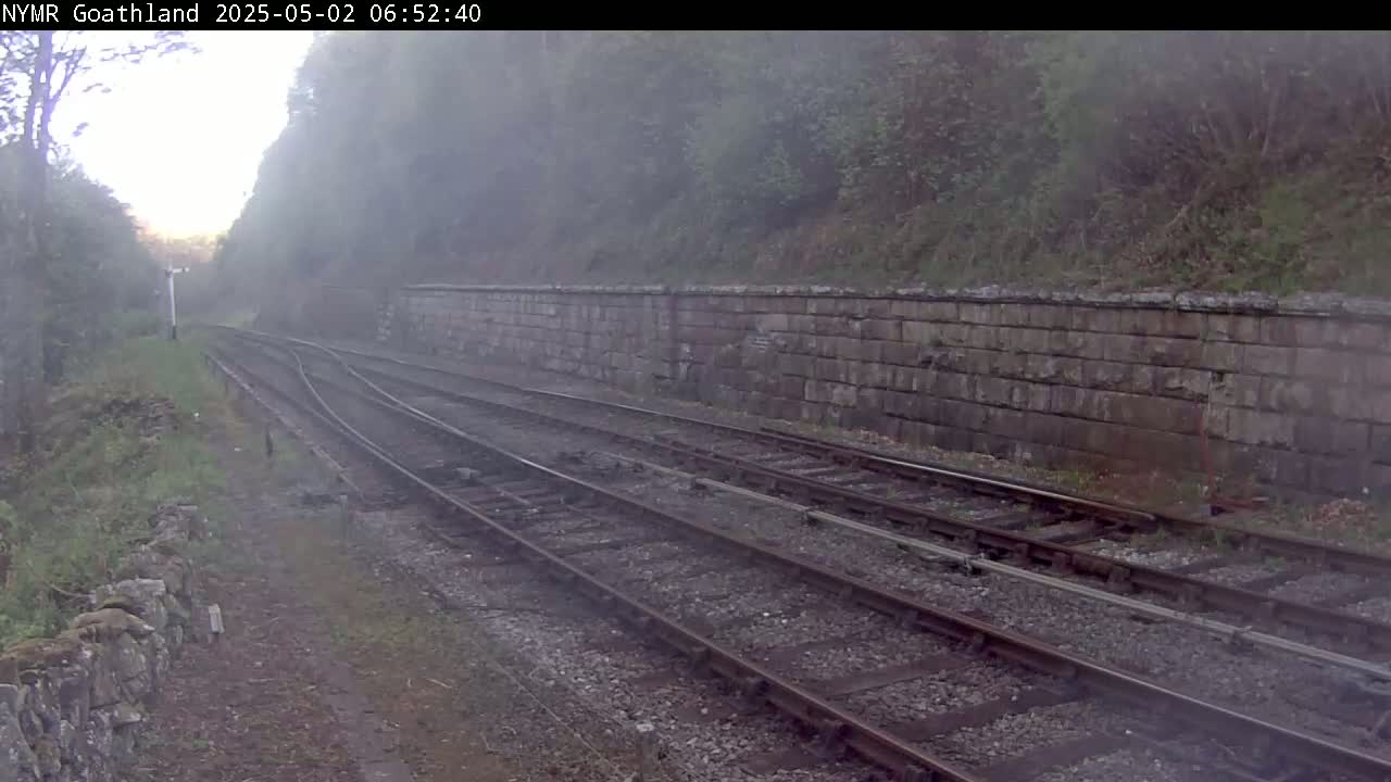 A foggy view of a railway track, running alongside a stone wall and bordered by vegetation.