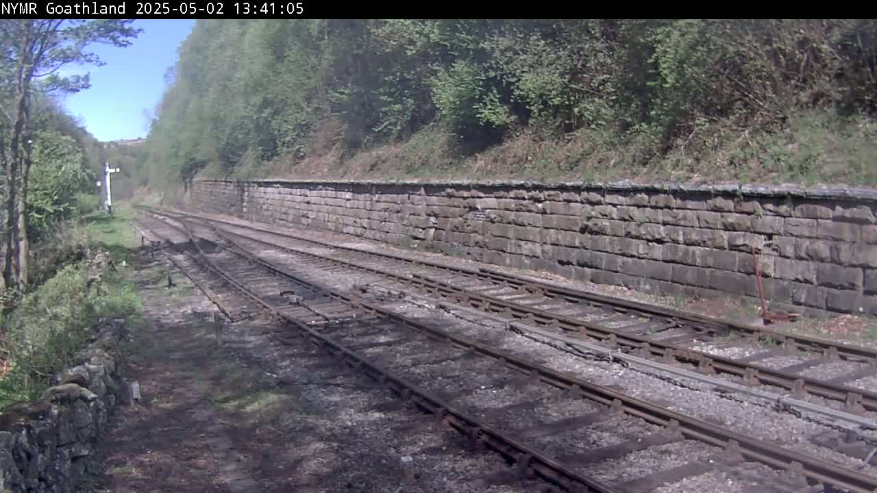 A view of a railway track on a sunny day, running alongside a stone wall and bordered by lush green vegetation.