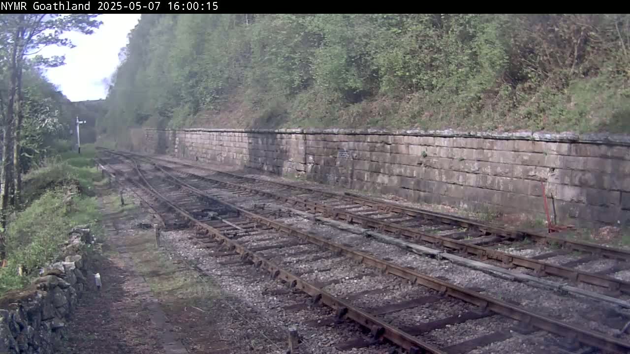 A view of railway tracks running alongside a stone wall, bordered by green vegetation on an overcast day.