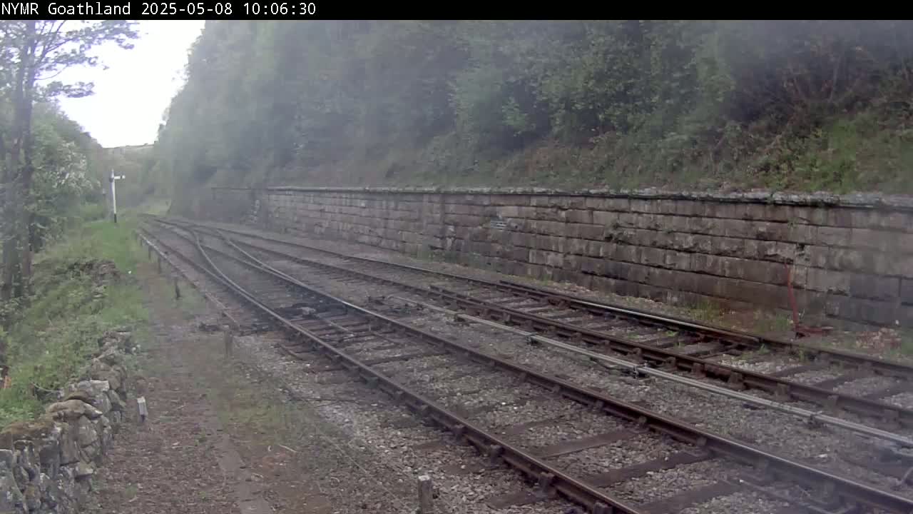 A misty view of a railway track diverging alongside a stone retaining wall, bordered by green vegetation.